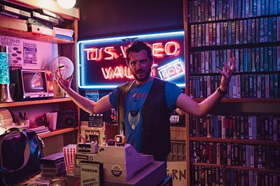 TJ (Kevin McGahern) with his arms raised in a video shop with an LED sign behind him saying 'TJ's Video Vault'