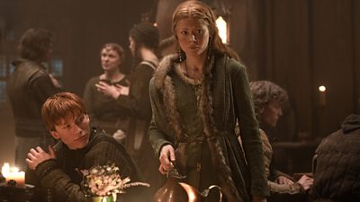 A woman wearing warm clothing from the Medieval period stands beside a banquet table holding a jug. She looks at something offscreen with a concerned expression. Characters in the background socialise. 