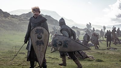 A man wearing chainmail and military clothing from the Medieval period stands on a battlefield. Holding a large wooden shield and a sword, he looks at something offscreen. Soldiers in the background continue the battle. 