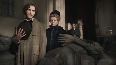 A man and woman in formal clothing from the Victorian-era stand in a large church beside a stone tomb. The man holds a bowler hat to his chest respectfully. A man in the background watches them. 