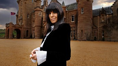 Claudia Winkleman is pictured outside the Traitors castle in daylight. She wears a black jacket over a white ruffled shirt.