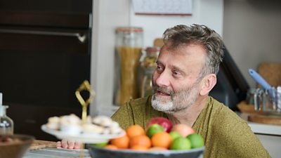 A man crouches beside a kitchen counter. A fruit bowl and a plate of mince pies sit on the work surface in front of him. 