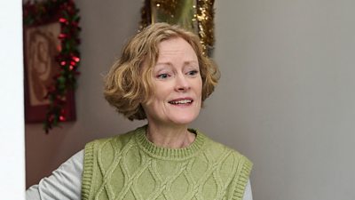 A woman holds open the front door to a house. The hallway behind her is decorated with tinsel. 