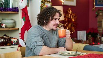 A man sits at a family dining table and holds a mug of tea. Christmas decorations litter the shelves behind him. A plate with leftover food sits on the table in front of him. 