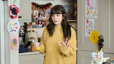 A young woman stands in a family kitchen. Her hands are outstretched in concern as she reacts to something happening offscreen. Children’s drawings are hung on the walls in the background alongside Christmas decorations. 