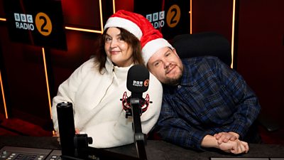 Ruth Jones and James Corden sit together behind the microphone in the ý Radio 2 studio wearing Santa hats. James rests his head on Ruth's shoulder.