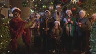 Kim conducts a choir of Christmas singers featuring Walford residents at the Christmas tree in the Square