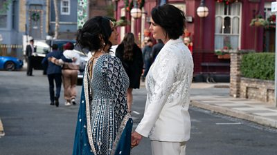 Suki and Eve stand holding hands on the Square, dressed for their wedding