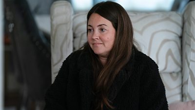 Stacey sits in an armchair looking at someone off screen