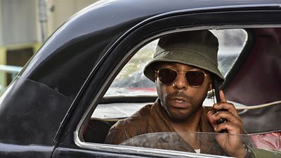 Don Gilet as DI Mervin Wilson, who sits in a car on his phone wearing a bucket hat and sunglasses