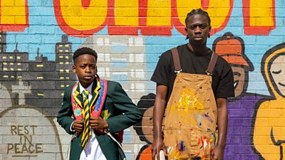 A young schoolboy in uniform stands next to an older teenager in an artist apron in front of a graffitied wall