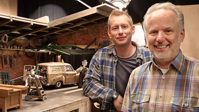 Two men smile to the camera in a behind-the-scenes photo. They stand beside a set for Wallace and Gromit, featuring clay models of the two iconic characters working on a van. 