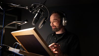 Behind-the-scenes photo from the audio recording of Wallace & Gromit: Vengeance Most Fowl. A man stands in front of a microphone and a script. 