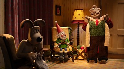 Gromit, Norbot, and Wallace in a sitting room. Wallace holds up a full-length knitted outfit and smiles. Norbot and Gromit sit in armchairs with knitting needles. 