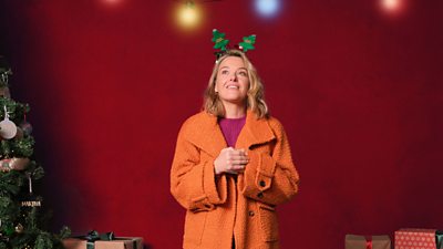 Sally Bretton stands against a red background wears an orange coat and a headband with two Christmas trees on it