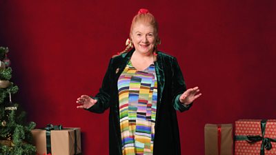 Felicity Montagu stands against a red background wearing a brightly coloured dress and a velvet green coat