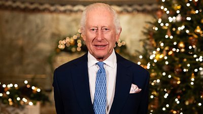 A photo of HM King Charles III stood in front of a Christmas tree glowing with golden lights.