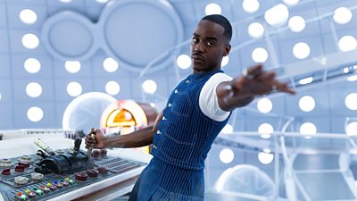 The Doctor, wearing a dark blue pinstripe waistcoat over a white t-shirt, leans back from the TARDIS console with one hand on a lever and the other reaching out towards something off camera