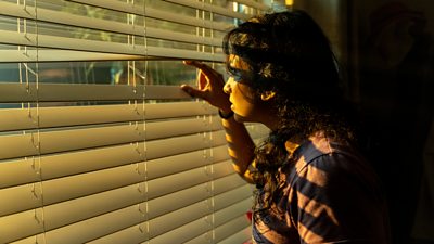 Belinda looks out a window through blinds