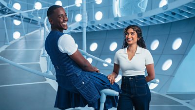 The Doctor and Belinda in the TARDIS. The Doctor sits on a rail, smiling, while Belinda stands by his side, resting a hand on his knee.