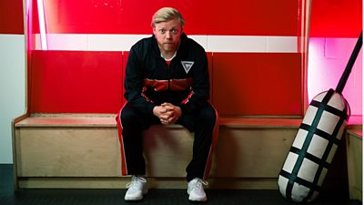 Rob Beckett sits on a light brown wooden bench in a changing room, wearing a tracksuit