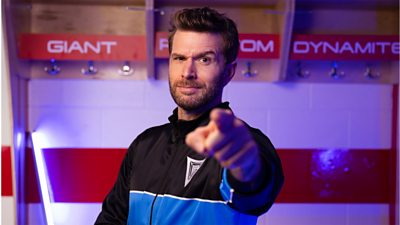 Joel Dommett points towards the camera with an eyebrow raised, standing in the Gladiators' changing room