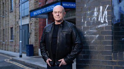 Ross Kemp in character as Grant Mitchell. He stands by a wall outside Walford East tube station, wearing a black jacket over a dark top.