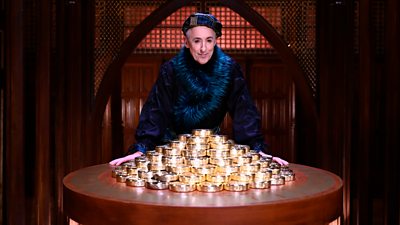 Alan Cumming stands in the castle behind a table piled high with discs of gold