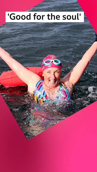 A smiling woman, Julie Wilson Nimmo, in the sea with her hands stretching to the sky wearing a pink swim hat and goggles