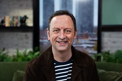 Matt is sat down on a green sofa smiling at the camera. He wears a stripy blue and white top with a brown velvet jacket over it.