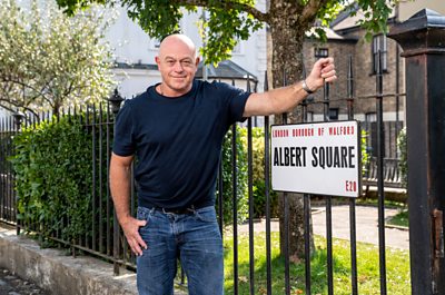 Ross stands smiling on Albert Square next to the street sign holding the park's railings. He wears a navy tshirt and jeans. Arthur's bench can be seen in the background.