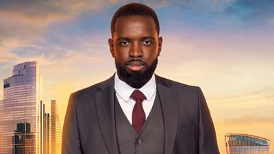 Frederick wears a grey suit and waistcoat with maroon red tie and white shirt. He has a full beard and short hair. A backdrop of the London skyline at sunset