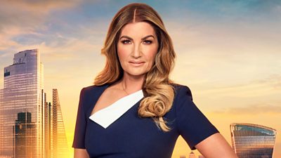 Karren Brady stands with her hand on hip in front of sunset London Skyline backdrop. She wears a navy dress with a white asymmetric collar