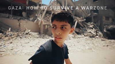 A young boy looks over his shoulder as he walks along a street of severely damaged buildings. Rubble spills from the buildings onto the road. 