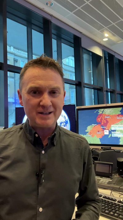 Matt Taylor wearing a grey shirt talks to the camera, with a computer showing a weather forecast in the background