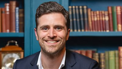 A man in a dark suit and white shirt smiles for the camera against the backdrop of a bookcase