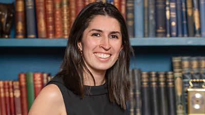 A woman with shoulder length dark hair wearing a black top stands smiling in front of a bookcase