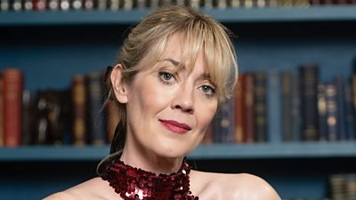 A woman with blonde hair tied back stands in front of a bookcase wearing a red sequin halterneck dress