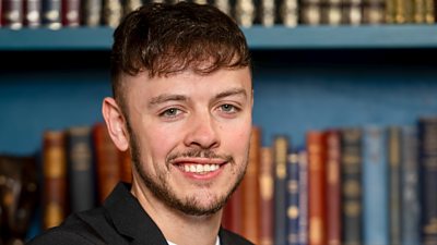 A man with brown hair and a short beard and moustache stands in front of a bookcase wearing a white t-shirt beneath a black suit jacket