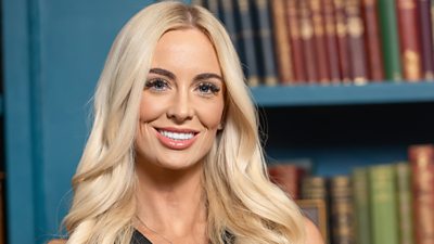 A woman with long blonde curled hair stands in front of a bookcase wearing a black dress