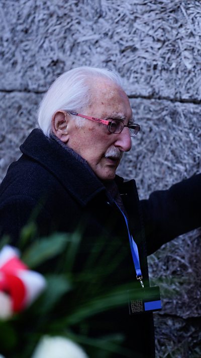 Elderly man rests hand on wall of death. Wreath can be seen in the foreground