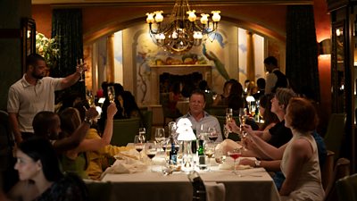 A large group of people sit around a dinner table in a restaurant raising their glasses in a toast to a man who sits at the head of the table. The room is dimly lit and a chandelier hangs from the ceiling.