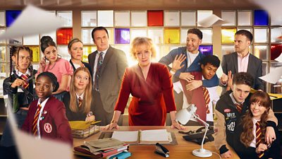 A group of students and teachers are gathered around a messy desk, where a woman at the centre stands with her hands resting on the desk. Chaos unfolds around her.