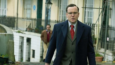 Joshua McGuire in character as Douglas Baxter, walking up a street wearing a three piece suit and glasses