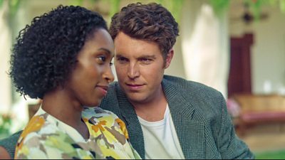 Tom Hughes stares intently at Rochelle Neil. Both have a half smile on their face and are sat down. Tom's arm is around Rochelle's