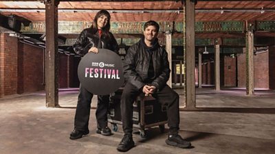 Deb Grant and Tom Ravenscroft inside an empty Victoria Warehouse. Tom is sat on a flight case and Deb is holding a 6  Music Festival roundel 