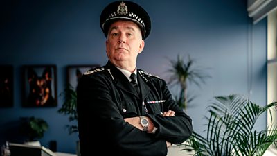 Jack Docherty in character as The Chief stood in police uniform with his arms folded.