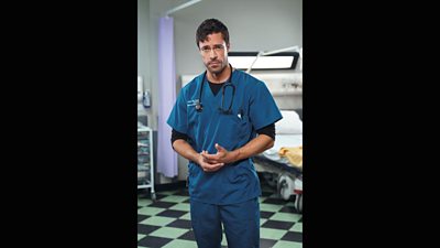 Olly Rix as Flynn Byron. He is wearing his hospital scrubs and standing on the ward whilst holding his hands and looking at the camera.