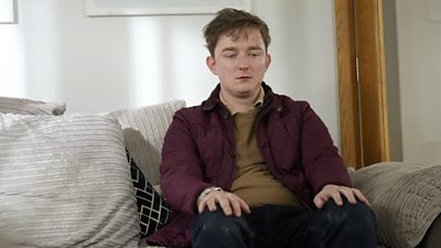 Callum sits on a sofa with his hands on his knees. He is wearing a maroon coat and looks forlorn 