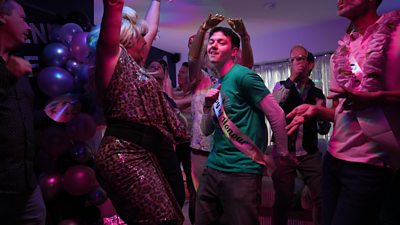 Jordan dances in a room full of people and balloons. Lit by pink lights, he wears a sash that reads boy of honour.